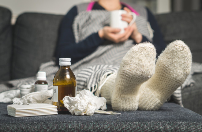  sick woman drinking hot beverage to ease flu symptoms, fever and virus. Dirty paper towels, cough medicine, and tissues on table. Ill person 